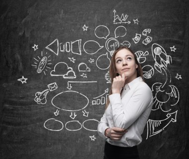 Young beautiful business woman is thinking about optimisation of social media process. Chalk icons are drawn on the blackboard.