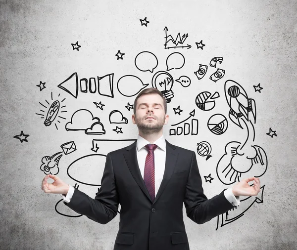 Meditating businessman in a suit. A concept of searching the best business development concept. Drawn business icons on the concrete wall. — Stok fotoğraf