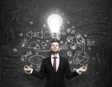Meditative young businessman is looking for new business ideas. Drawn business icons on the black chalk board and a light bulb as a concept of new idea.