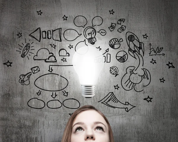 Young business lady is looking for new business ideas. Drawn business icons on the concrete wall and a light bulb as a concept of new idea. — Stok fotoğraf