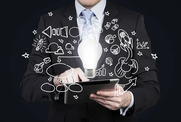 Businessman is looking for new business ideas in tablet. Flying business icons and a light bulb as a concept of new idea. Black chalk board on the background. — Stockfoto