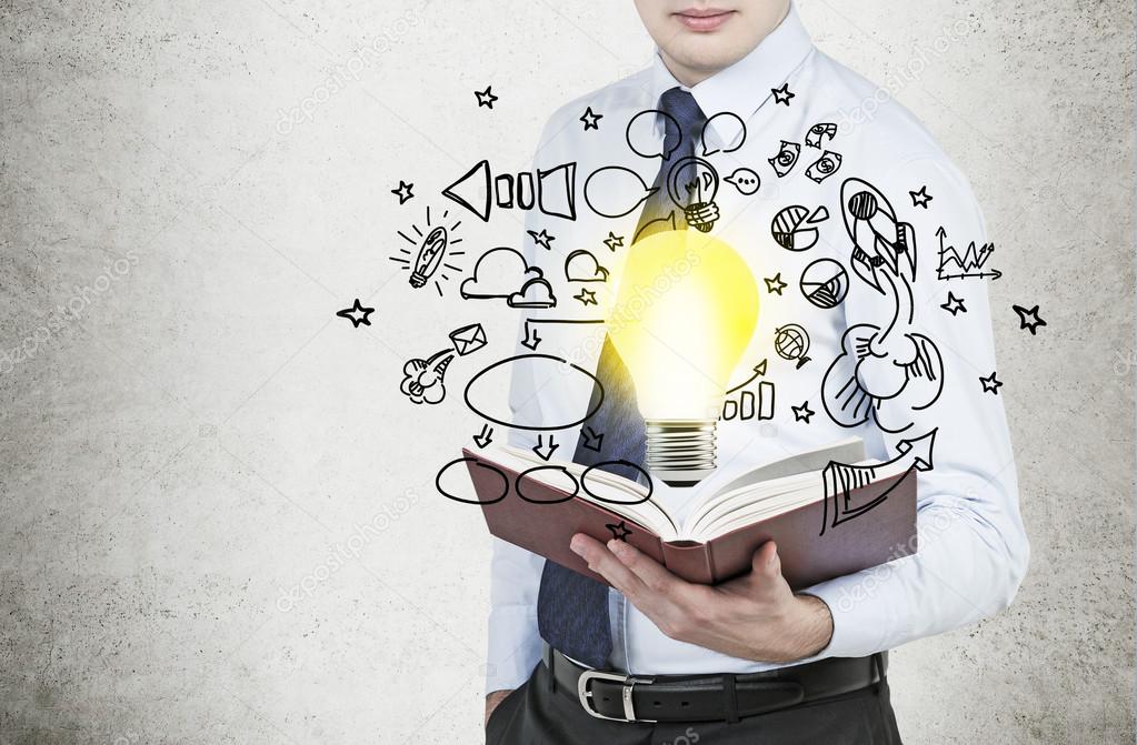 Young businessman is holding a book with flying around business icons and a light bulb as a concept of the new business ideas.