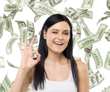 Brunette woman shows ok sign. Dollar notes are falling down over isolated background.