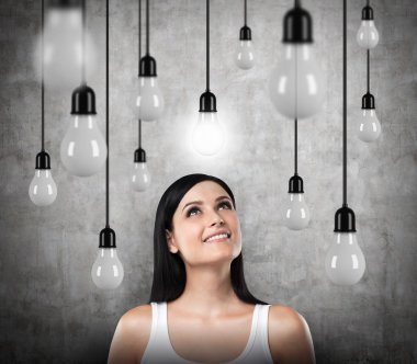A dreamy brunette looks upward. Lots of light bulbs, one of them is turned on. Concrete background.