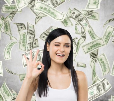 Brunette woman shows ok sign. Dollar notes are falling down over concrete background.