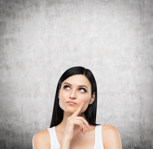 A portrait of a pensive brunette lady in a white tank top. Concrete background.