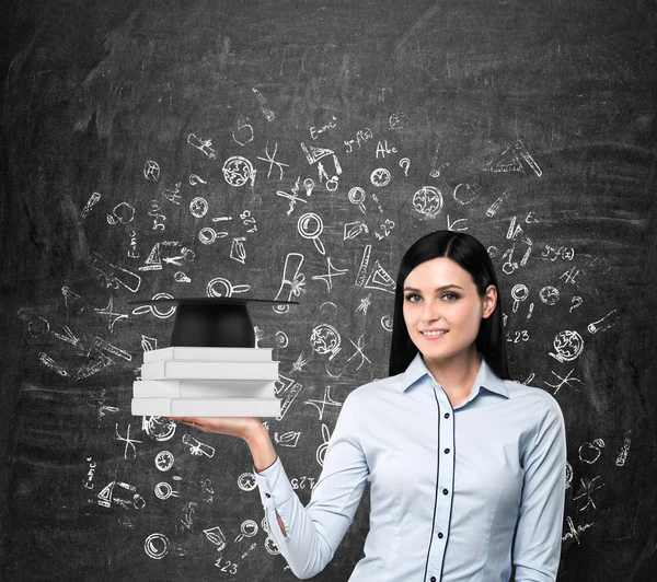 Brunette lady holds books with a graduation hat. Educational icons are drawn over the black chalk board. — Stock Fotó