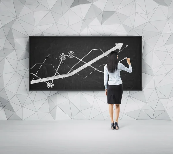 Rear view of full-length brunette woman who is presenting growing line graph on the black board. — Stockfoto