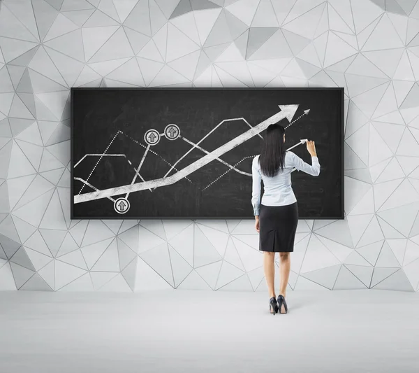 Rear view of full-length brunette woman who is presenting growing line graph on the black board. — Stock Fotó