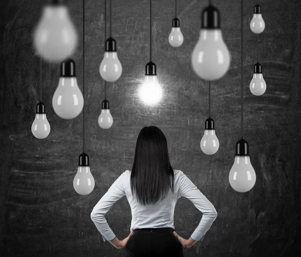 Rückansicht der brünetten Dame, die auf die hängenden Glühbirnen blickt. ein Konzept der Suche nach neuen Ideen. schwarze Kreidetafel Hintergrund. — Stockfoto