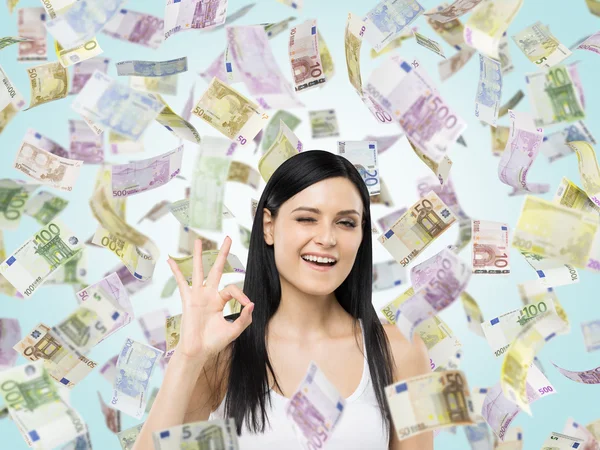 Brunette woman shows ok sign. Euro notes are falling down over blue background. — стокове фото