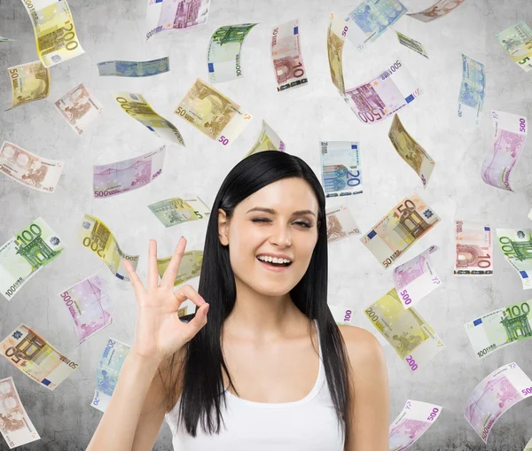 Brunette woman shows ok sign. Euro notes are falling down over concrete background. — Stockfoto