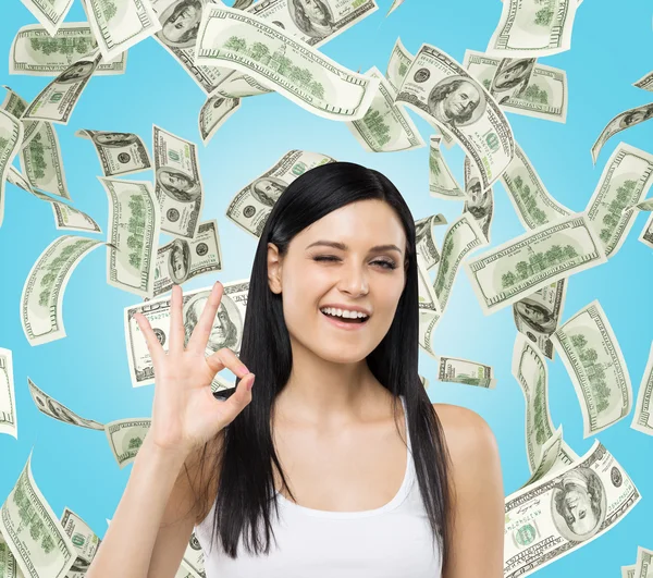 Brunette woman shows ok sign. Dollar notes are falling down over blue background. — Stockfoto