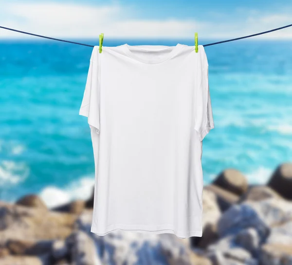 Primo piano di una t-shirt bianca sulla corda. Mare e rocce sfocate come sfondo . — Foto Stock