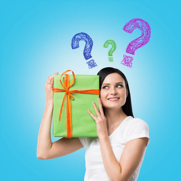Eine brünette Frau hält eine grüne Geschenkschachtel mit Fragezeichen als Konzept der Geschenkunsicherheit in der Hand. blauer Hintergrund . — Stockfoto