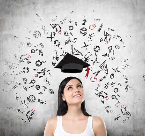 A portrait of a dreamy brunette lady about entering into the university. Concrete background with drawn educational icons and flying graduation hat. — Zdjęcie stockowe