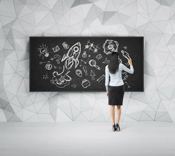 Rear view of full-length brunette woman who is drawing business icons on the black board. — Stock fotografie