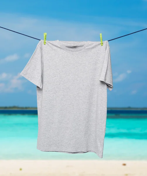 Primo piano di una t-shirt grigia sulla corda. Spiaggia di sabbia tropicale e oceano come sfondo . — Foto Stock