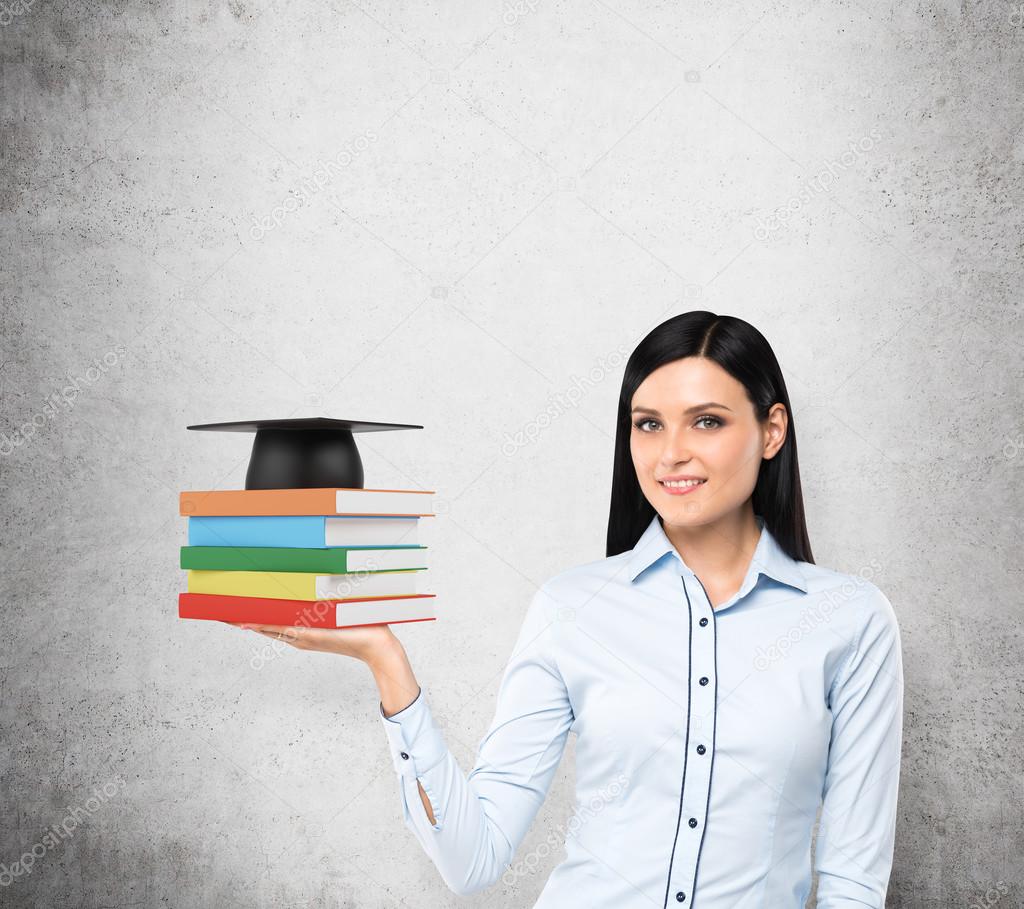 A portrait of a brunette lady with the open palm who is holding colourful books and a graduation hat. A concept of necessity of education. Concrete background..