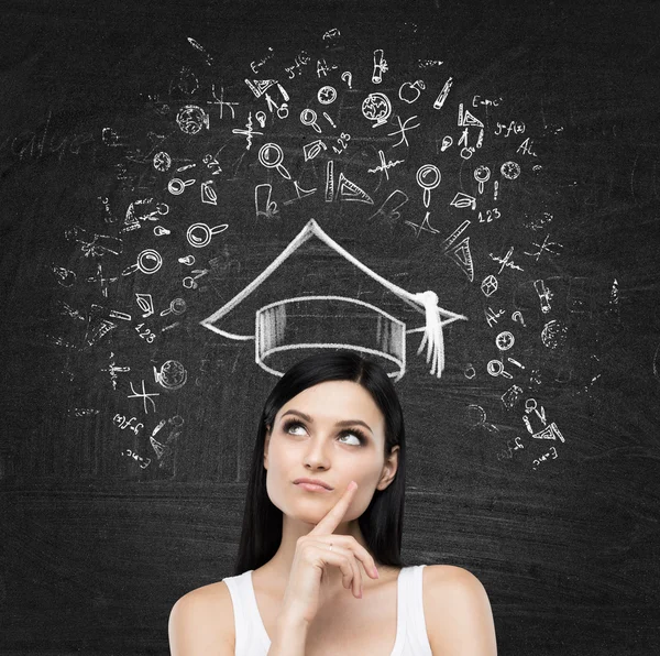 Young brunette lady is thinking about studying at the university. Educational icons are drawn on the black chalk board. — Stockfoto