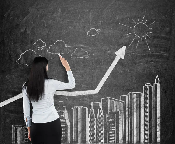Geschäftsfrau zeichnet Flussdiagramm von Wolkenkratzern auf schwarze Kreidetafel. — Stockfoto