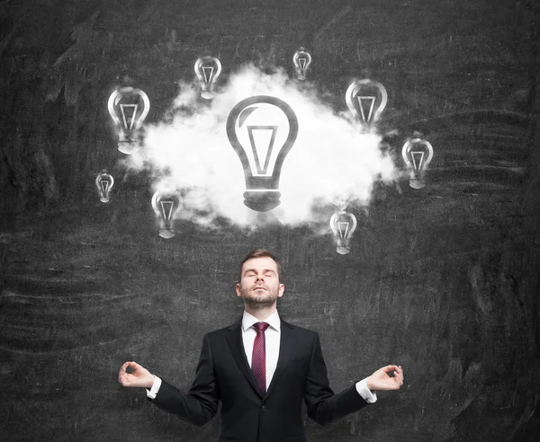 Meditative businessman at the cloud with light bulbs as a concept of the new business ideas. Black chalk board as a background. — Stok fotoğraf