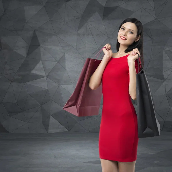 Beautiful brunette woman in a red dress is holding fancy shopping bags. Contemporary background. — Stockfoto