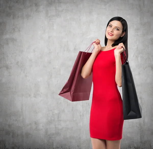 Vacker brunett kvinna i en röd klänning håller fancy shoppingkassar. Konkreta bakgrunden. — Stockfoto
