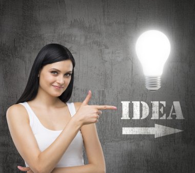 Brunette woman is in a white tank top. She points out the lightbulb and the word IDEA with the arrow. A concept of brainstorm. Dark concrete wall on the background.