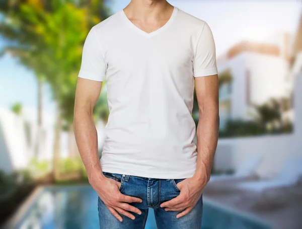 Young man in a white V shape t-shirt, hands in pockets. Villa with swimming pool in blur on the background. — 스톡 사진