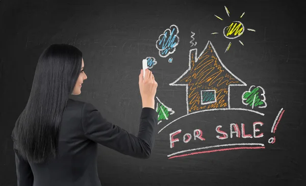 Brunette real estate agent is drawing a new colourful house on the black chalkboard. — Stock Photo, Image