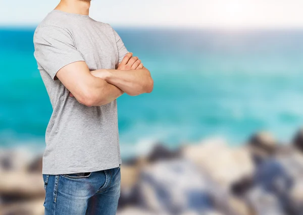 Vista lateral do jovem em uma camiseta cinza com as mãos cruzadas. Fundo vista mar em borrão . — Fotografia de Stock
