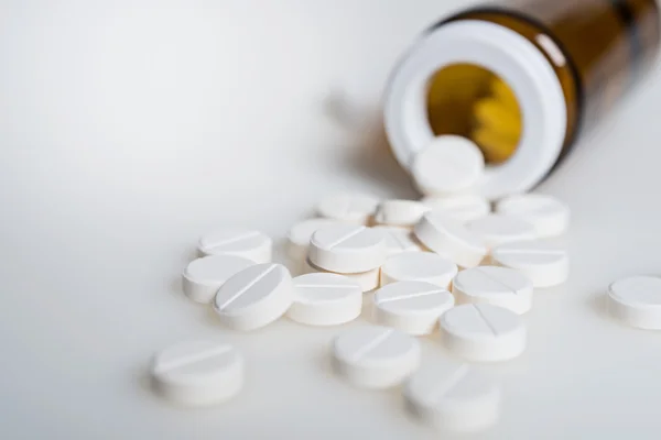 Garrafa médica de vidro e pílulas brancas que são derramadas na superfície branca — Fotografia de Stock