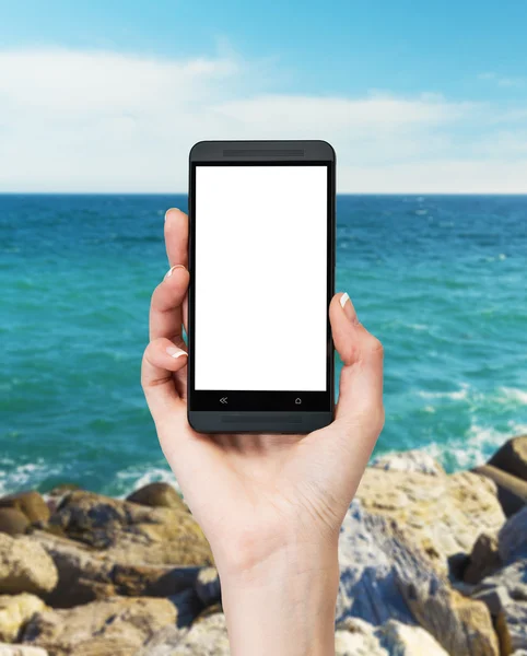 A hand is holding a smartphone with white copy space screen. Sea view on the background in blur. — Stockfoto
