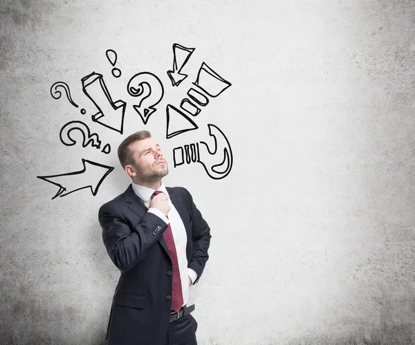 Thoughtful businessman is trying to find a right solution. Question marks and arrows are drawn on the concrete wall. — Stock fotografie