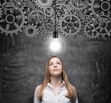 Young charming business woman is looking at the light bulb as a concept of innovative business ideas. Gears as a concept of the brainstorm. Black chalkboard as a background.