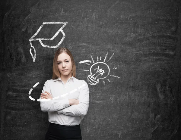 Beautiful young lady with crossed hands is thinking about education. A graduation hat and a light bulb are drawn on the chalkboard above the lady. — Stockfoto