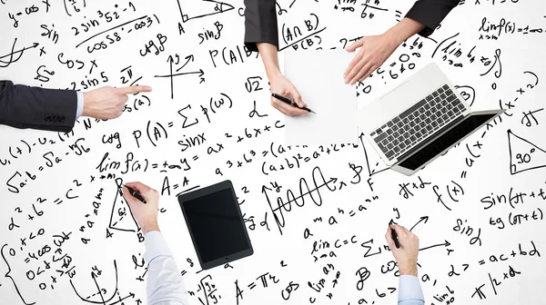 Eine Draufsicht auf einen Brainstorming-Prozess. Kollegen lösen Mathe-Aufgaben. Laptop, Tablet und mathematische Formeln werden über die Oberfläche auf dem Tisch gezeichnet. — Stockfoto