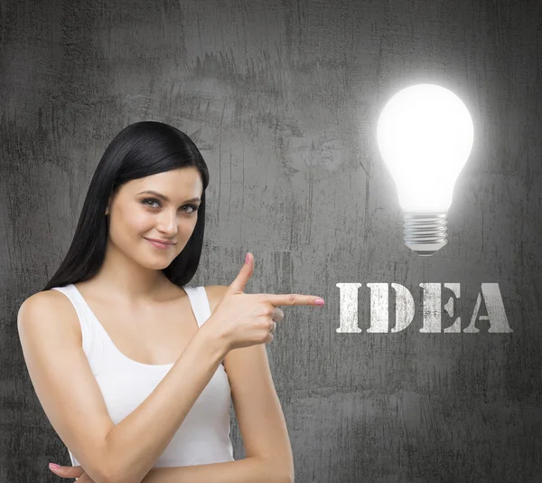 Brunette woman is in a white tank top. She points out the lightbulb and the word IDEA. A concept of brainstorm. Dark concrete wall on the background. — Stok fotoğraf