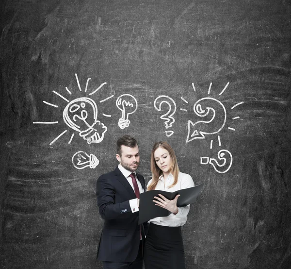 Business couple are arguing about business project . A couple in formal clothes are holding a black document folder. Light bulbs and question marks are drawn on the wall behind them. Black chalkboard — ストック写真