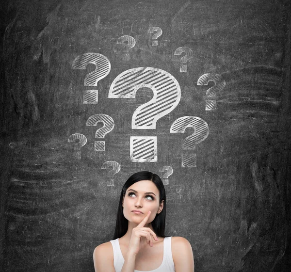 Thoughtful brunette lady is trying to find a proper solution. Question marks are drawn on the black chalkboard. — Stockfoto