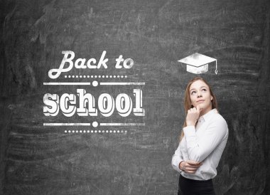 Young schoolgirl is thinking about future academic year. Words: ' back to school ' are written on the black chalkboard.