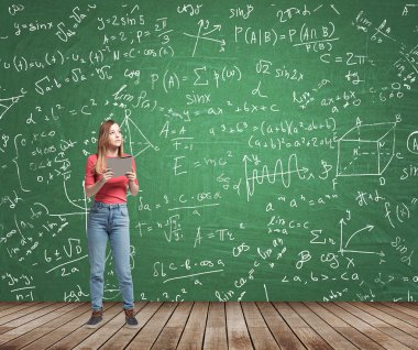 Young lady is pondering about complicated math problem. Formulas and graphs are drawn on the green chalkboard.