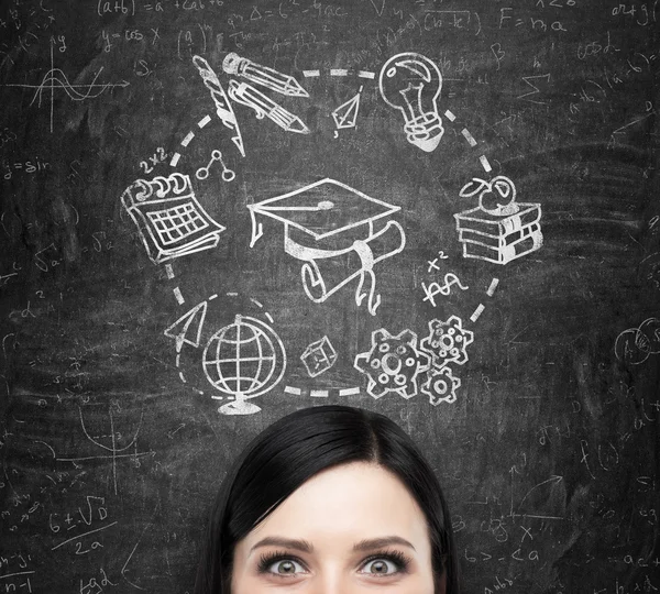 A forehead of the brunette lady who thinks about studying and graduation. Educational icons are drawn on the black chalkboard. — 图库照片