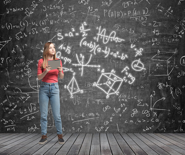 Young lady is pondering about complicated math problem. Formulas and graphs are drawn on the black chalk wall.