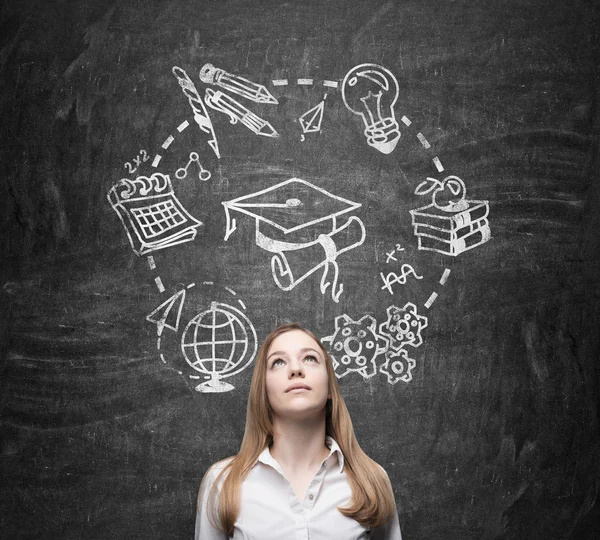 Young beautiful lady thinks about studying and graduation. Educational icons are drawn on the black chalkboard. — Stockfoto