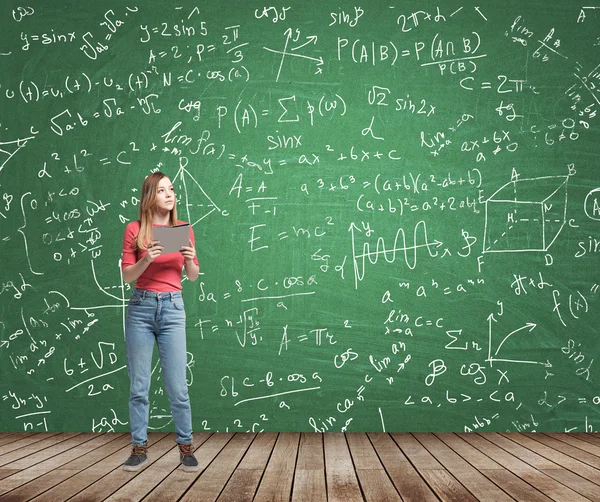 Young lady is pondering about complicated math problem. Formulas and graphs are drawn on the green chalkboard. — Stock Fotó