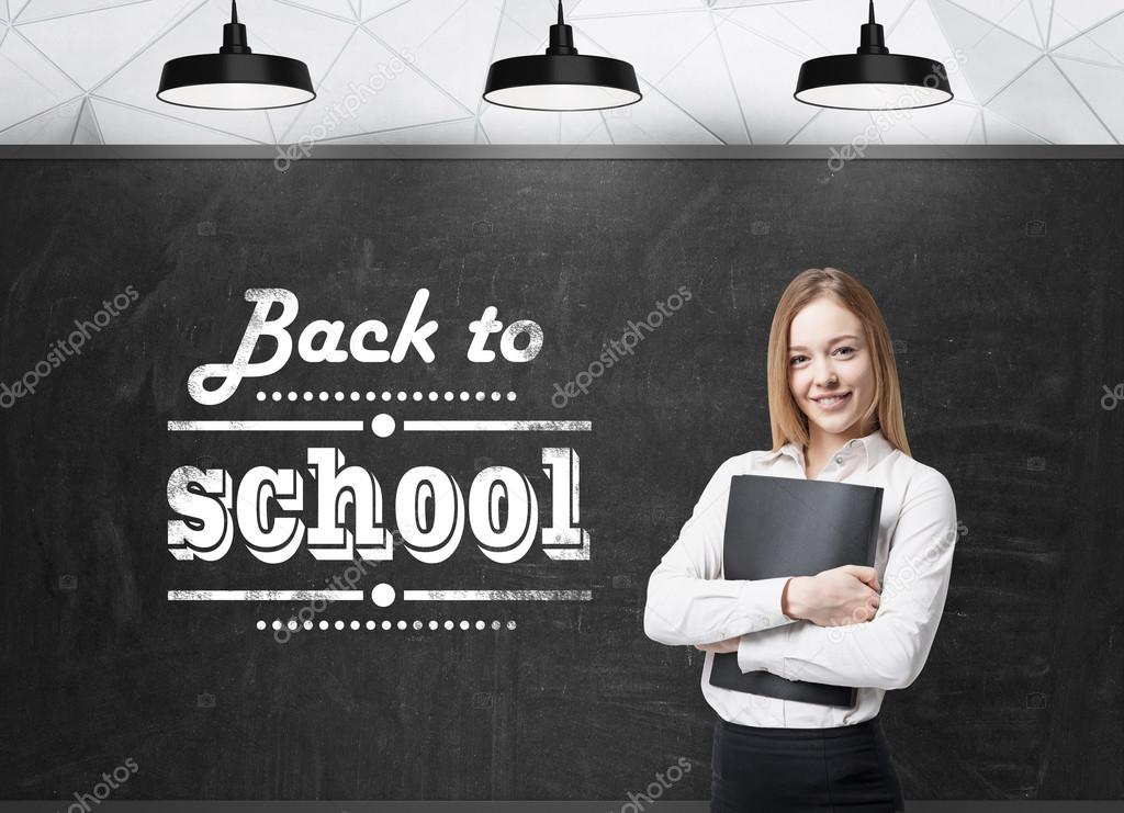 Young lady with a black folder is thinking about future academic year. Words: ' back to school ' are written on the black chalkboard.