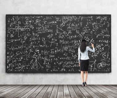 Full length of the rear view of the brunette girl who is writing down math formulas on the black chalkboard. A concept of university education.