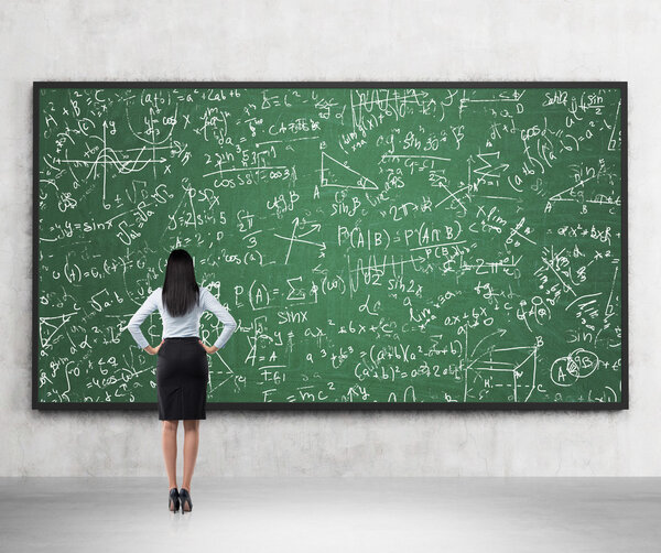 Full length of the rear view of the brunette lady who is looking at the math formulas which are drawn on the green chalkboard. A concept of university education.